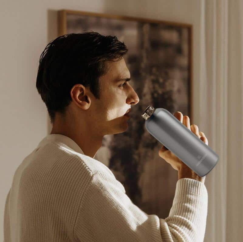 Man drinking from a gray metal bottle indoors while wearing a white sweater.