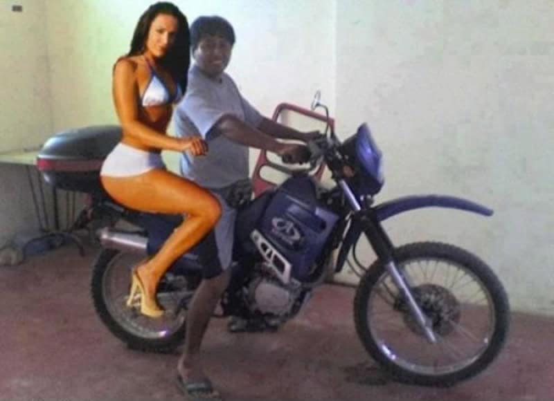 Woman in white swimsuit sitting behind man on a blue motorcycle in a garage setting.