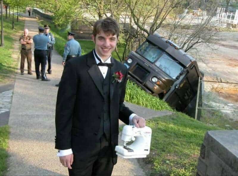 Man in tuxedo holding a white device, with a tilted vehicle in the background.