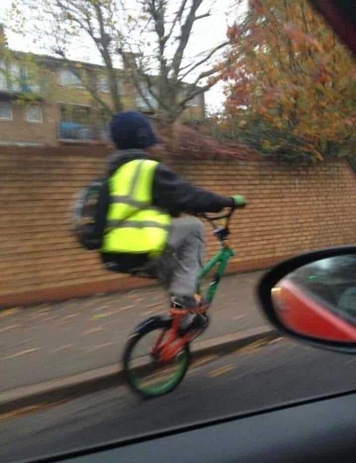 Child in a yellow vest riding a bike on the sidewalk, seen from car window.