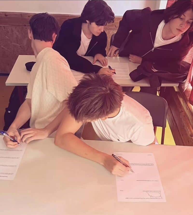 Four students working on papers at a desk, one leaning over, others sitting or standing.