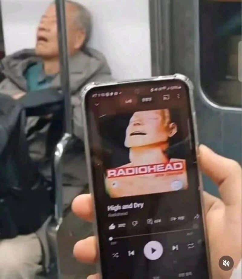 Person holding phone showing Radiohead album cover, with elderly man in background.