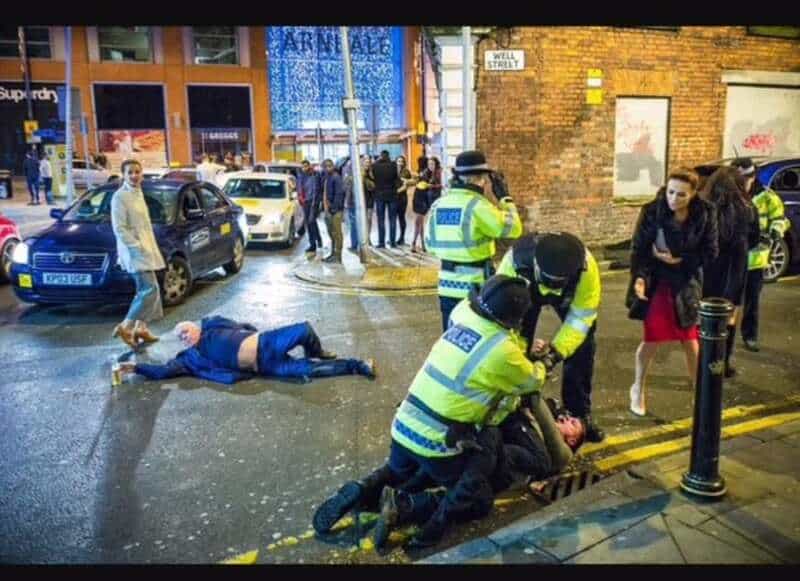 Police officers assist a person on the street at night, with a fallen man and onlookers nearby.
