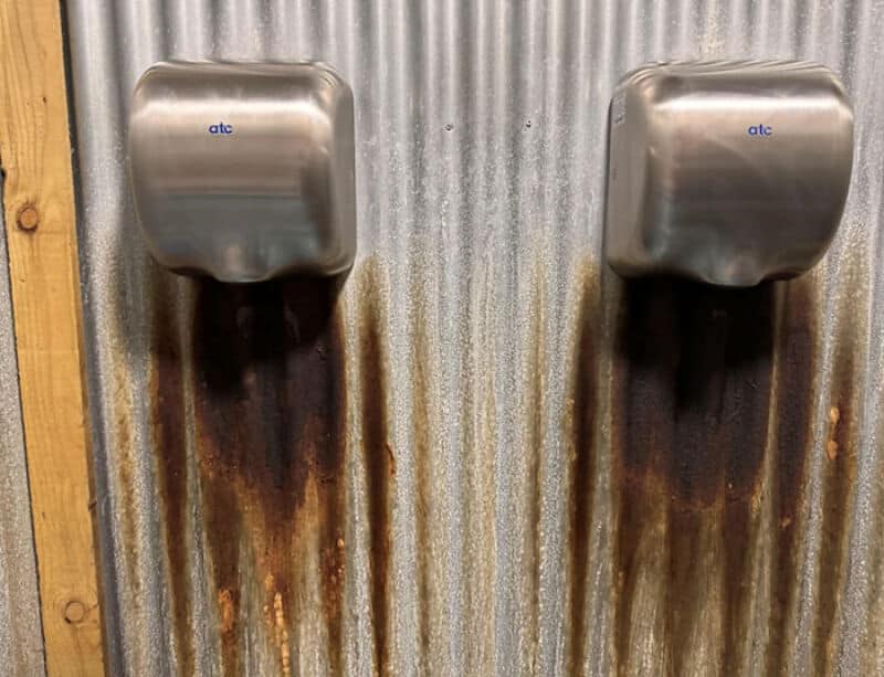 Two rusted metal hand dryers mounted on a corrugated metal wall in a bathroom.