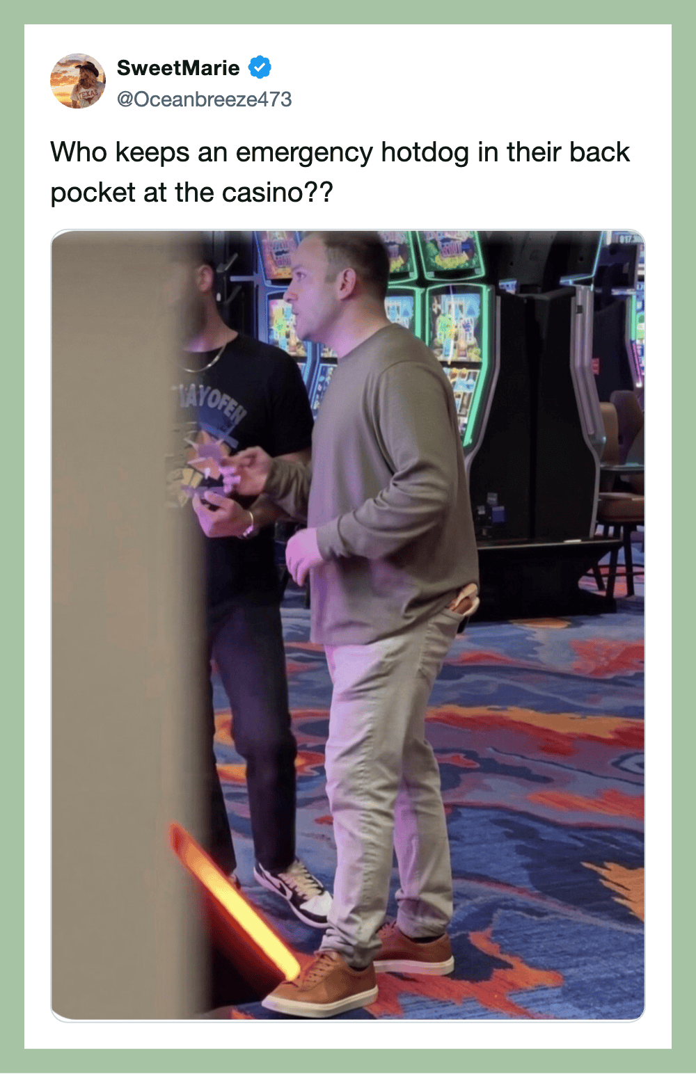 Man in gray hoodie holding a hotdog in a casino with slot machines in the background.