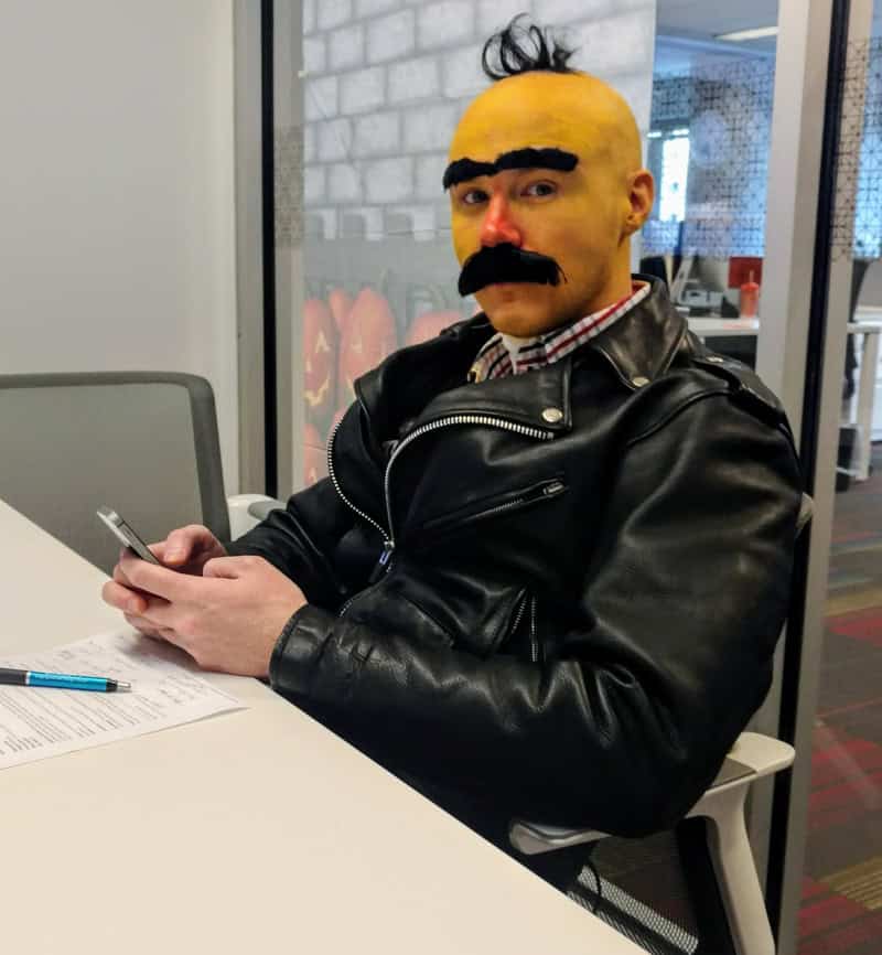 Person with yellow painted face, black eyebrows and mustache, wearing a black leather jacket, seated.