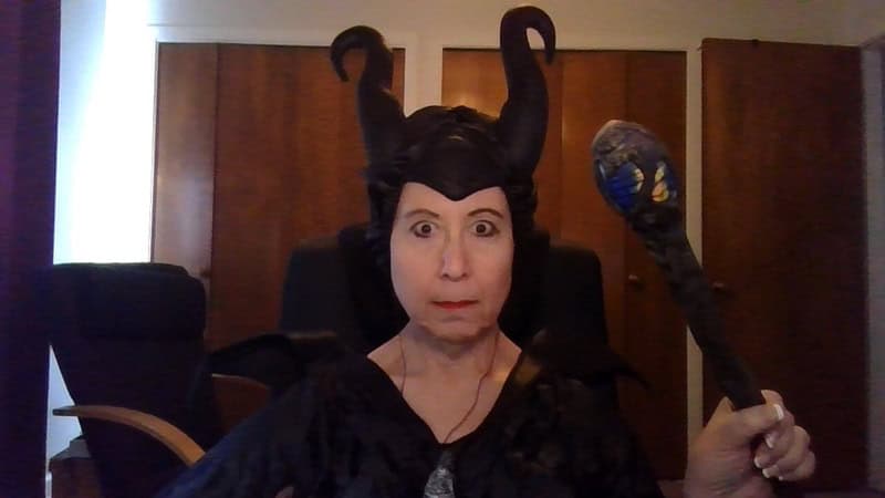 Woman in a black horned costume with staff, seated indoors.