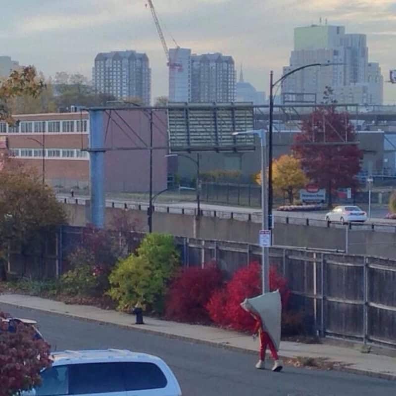 Person walking along a sidewalk with a large white cone-shaped object over their head, red shirt, in.