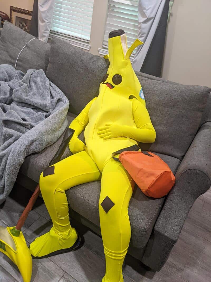 Person in a bright yellow banana costume sitting on a gray couch, holding a small orange bag.