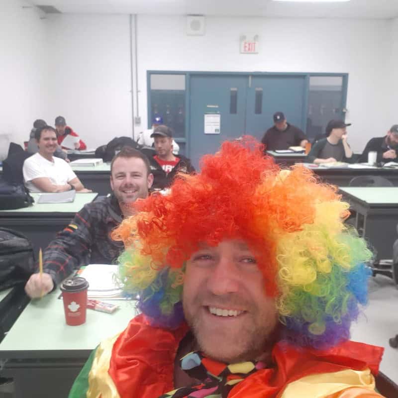 Man wearing a rainbow wig and colorful scarf smiles in a classroom with others at desks.