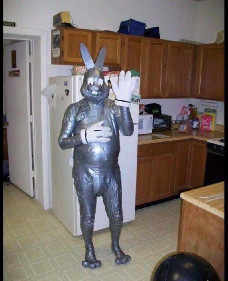Person in a full-body silver bunny costume waving in a kitchen.