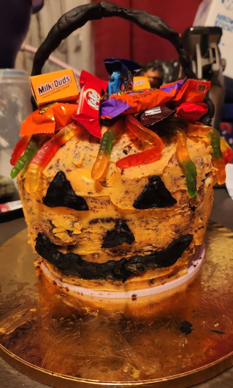 Halloween-themed cake with orange frosting, gummy worms, chocolate chips, and candy bars on top; a j.