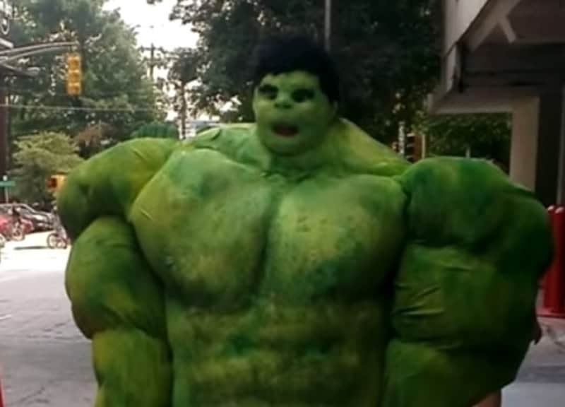Green inflatable Hulk costume walking on street with trees and buildings in background.
