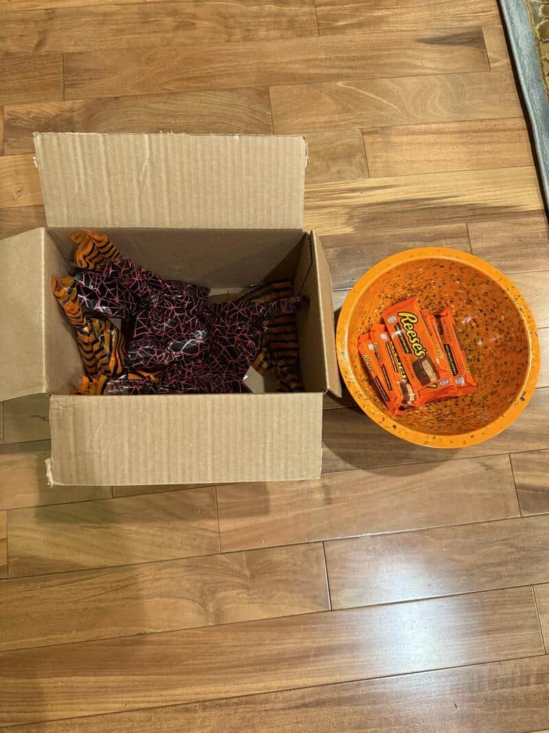 Halloween-themed box with costumes and a orange bowl of candy on wooden floor.