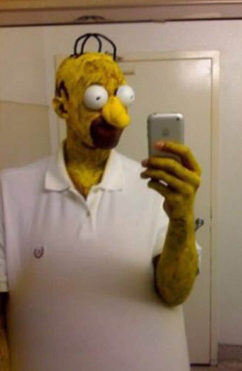 Homer Simpson-like character with yellow wig and white polo taking a mirror selfie.