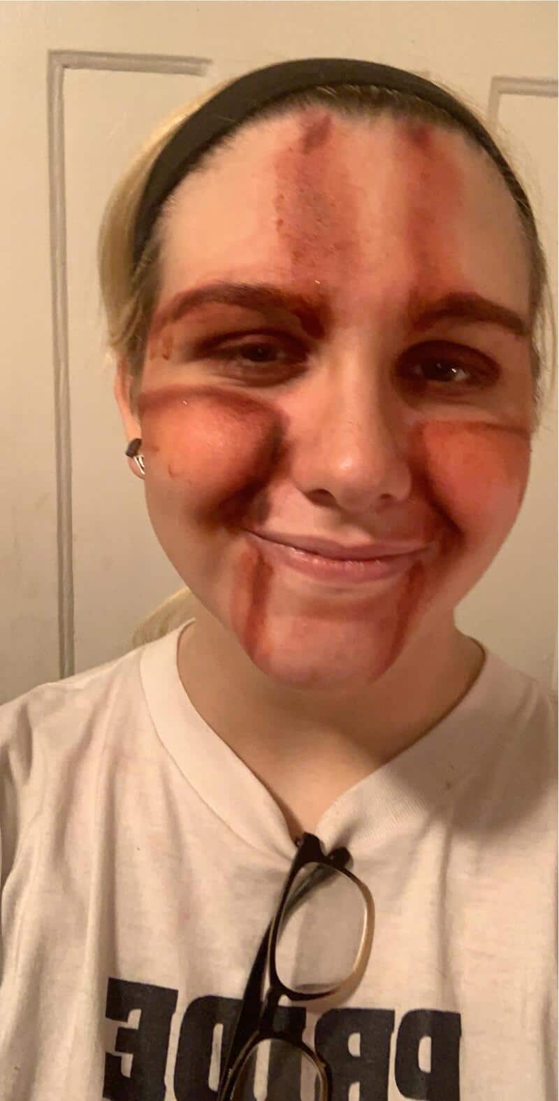 Person with makeup resembling a bloody mask, smiling, wearing white shirt and glasses hanging from a.