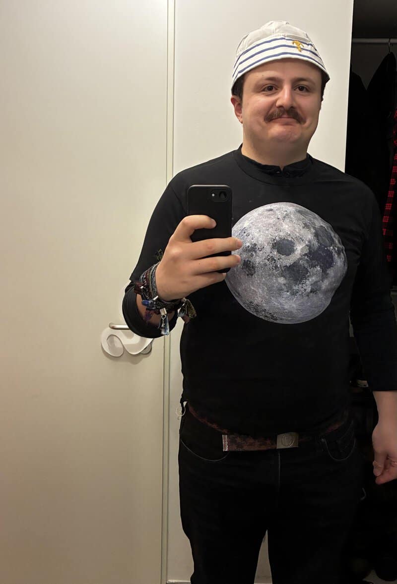 Person wearing a shirt with a large moon graphic taking a mirror selfie, white beanie, bracelets, in.