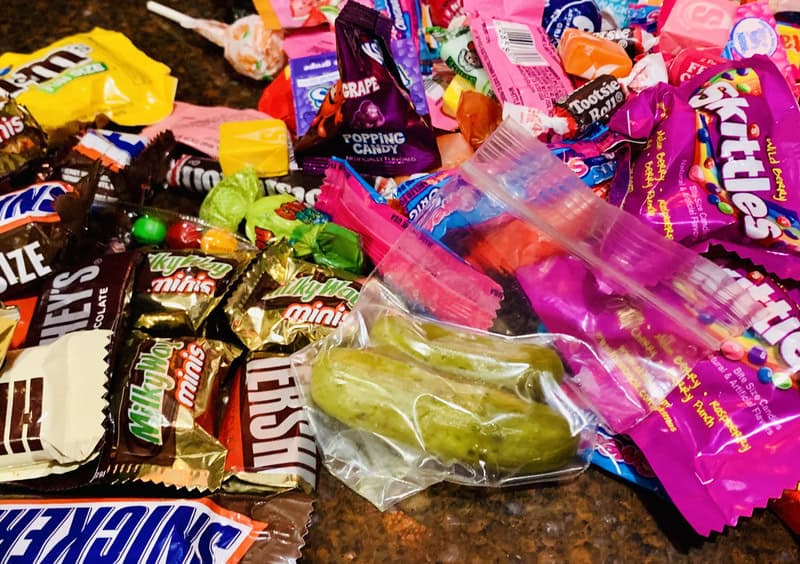 Assorted candy wrappers scattered with a single green pickle in a clear bag amid colorful snacks.