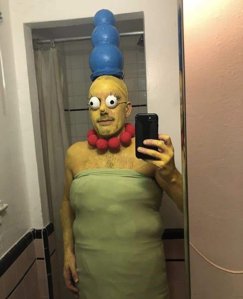 Person in a green dress with blue stacked hat, red necklace, large googly eyes, taking a mirrorself;.