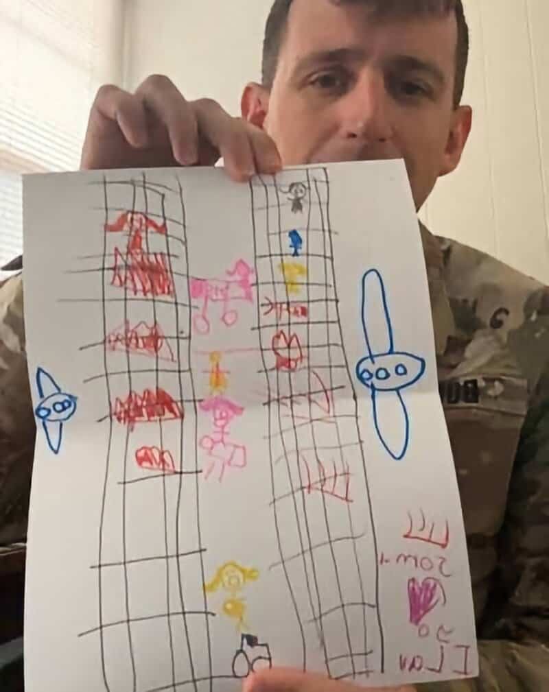 Man in military uniform holding a child's drawing on white paper with multiple colorful figures andy.