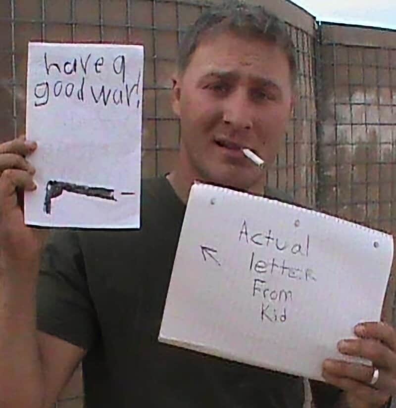 Man holding two signs: one says 'have a good war' with a drawing of a gun; the other reads 'Actual l.