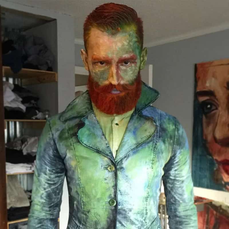 Man with green and red face paint and green coat in a room with artwork in the background.