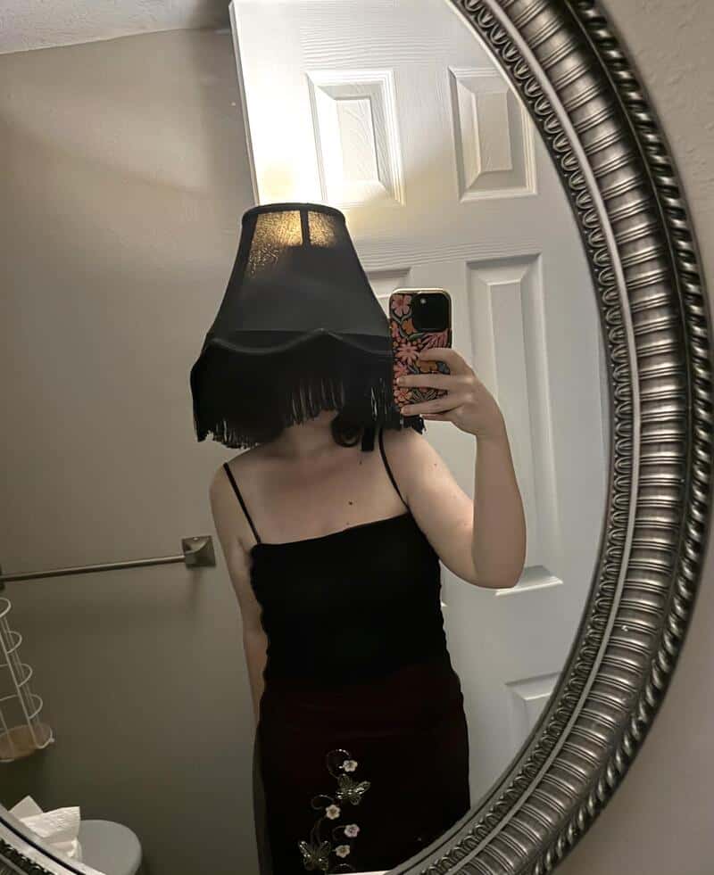 Person in a black sleeveless top and dark skirt taking a mirror selfie with a black lampshade over/遮.
