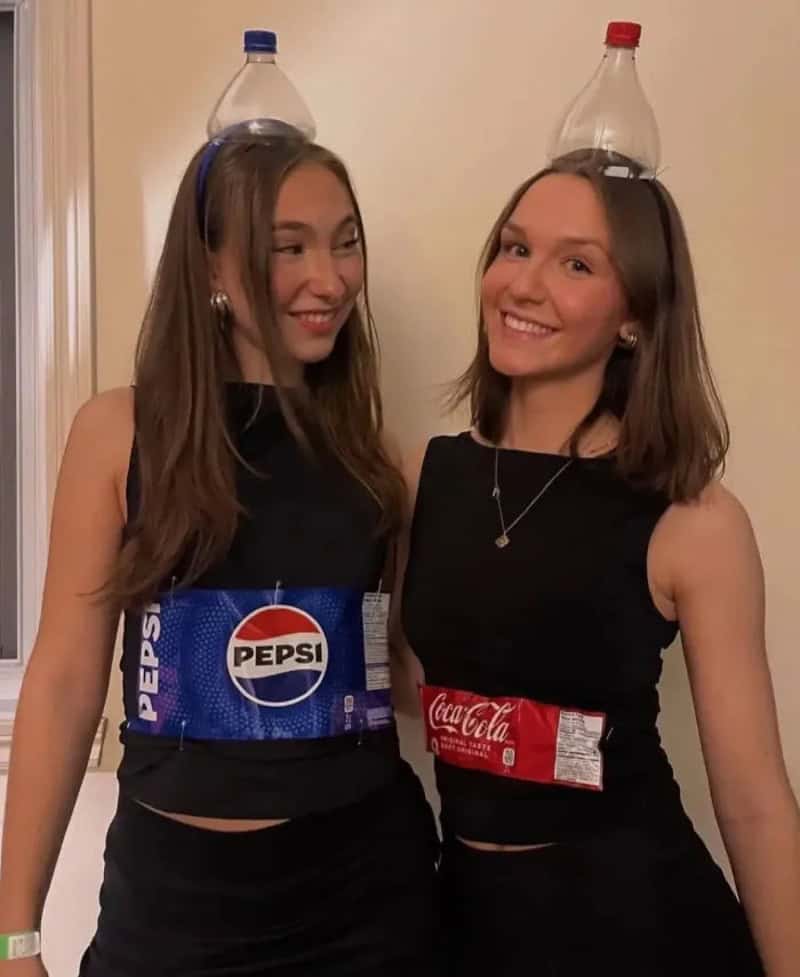 Two young women wearing black outfits with soda-pack accessories: Pepsi on the left and Coca-Cola on.