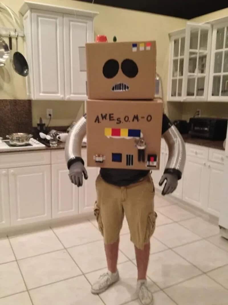 Person wearing a cardboard robot costume in a kitchen; box head with eyes and mouth, labeled Awesome.