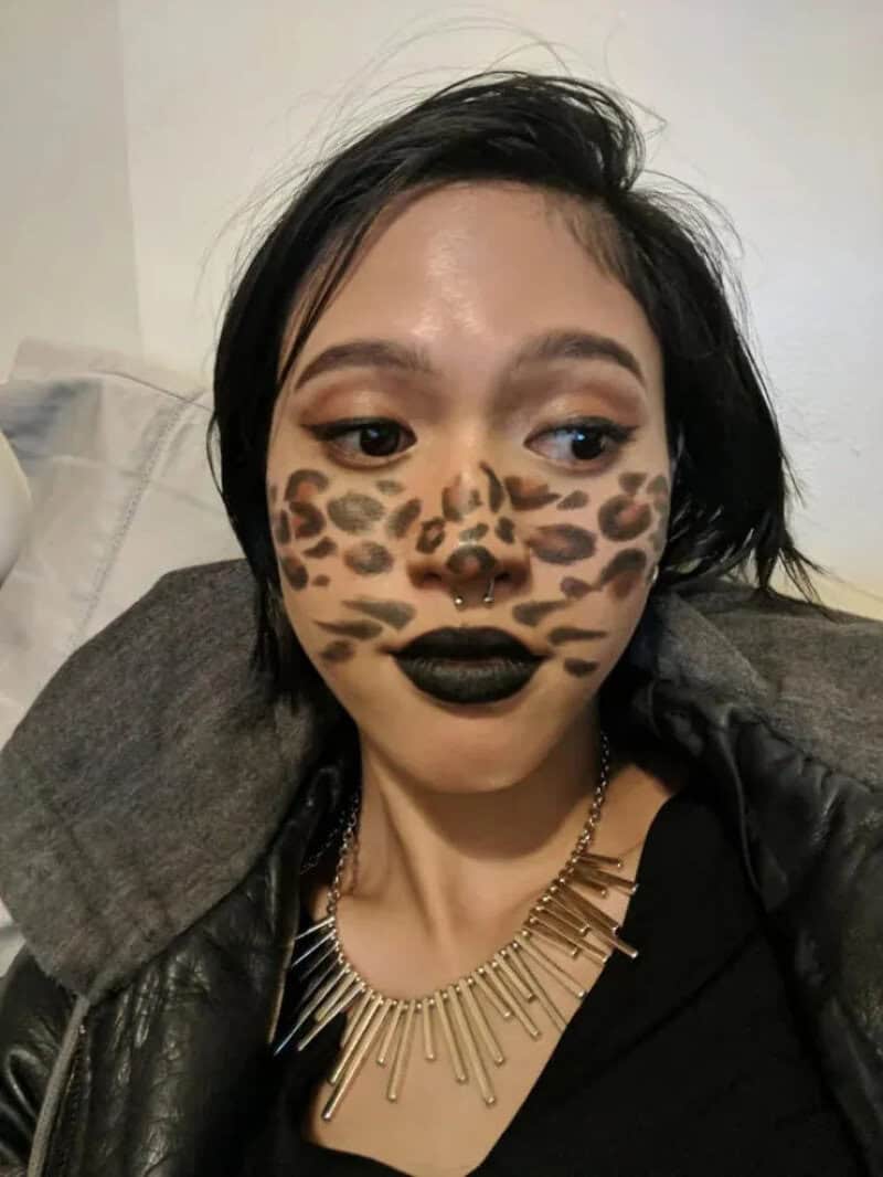 Person with leopard print face makeup, black lipstick, short dark hair, wearing a spiked necklace,,.