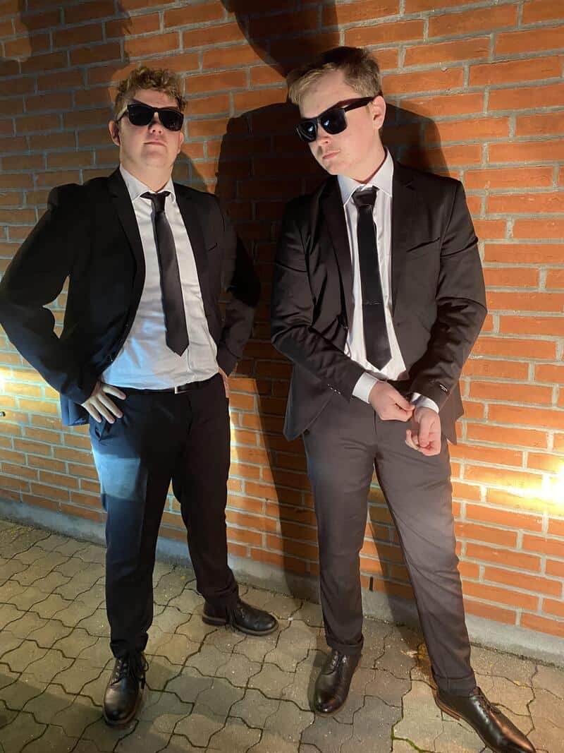 Two men in black suits and sunglasses standing against a brick wall; one has hands on hips, theother.