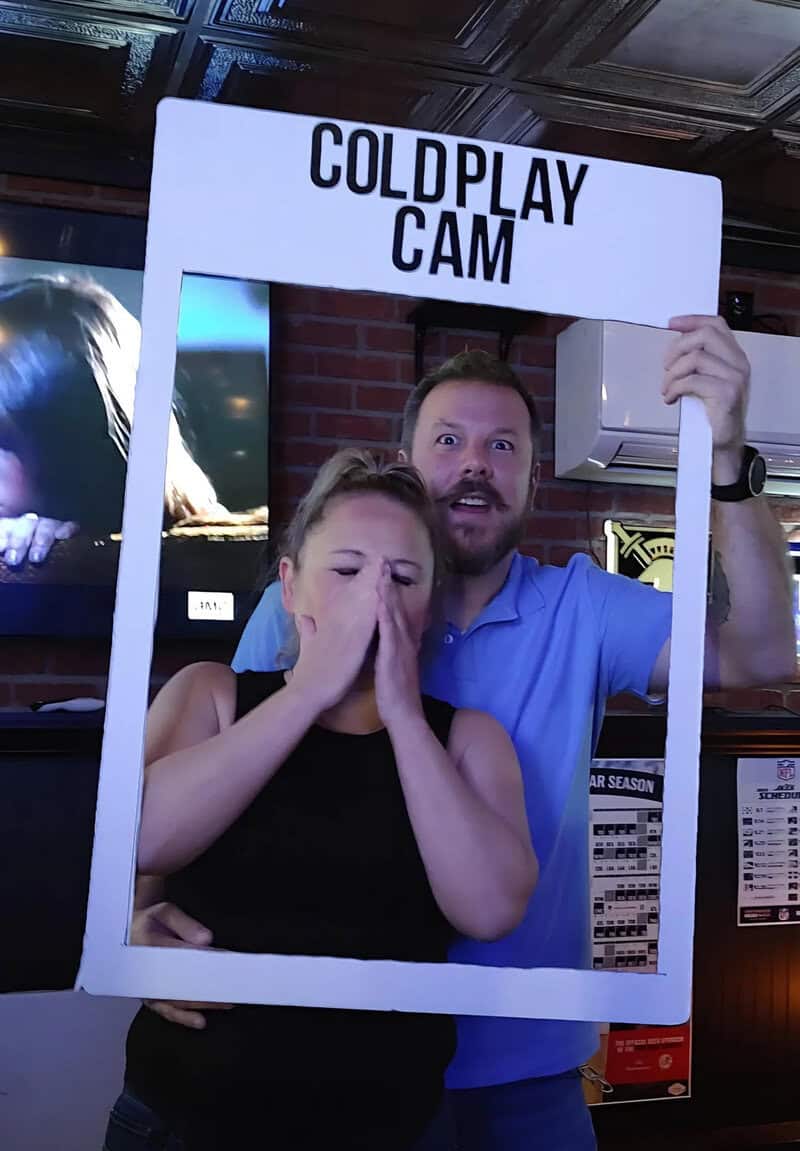 Coldplay Cam frame held by a man and a woman covering mouth, inside a venue.