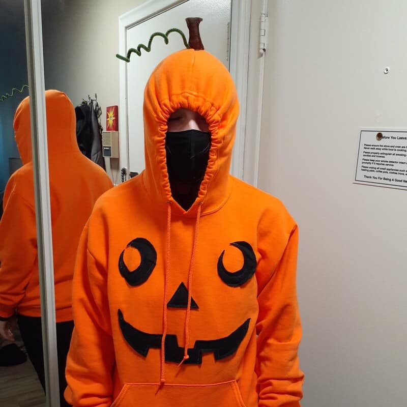 Person wearing orange pumpkin hoodie with jack-o'-lantern face and black mask in a mirror reflection.