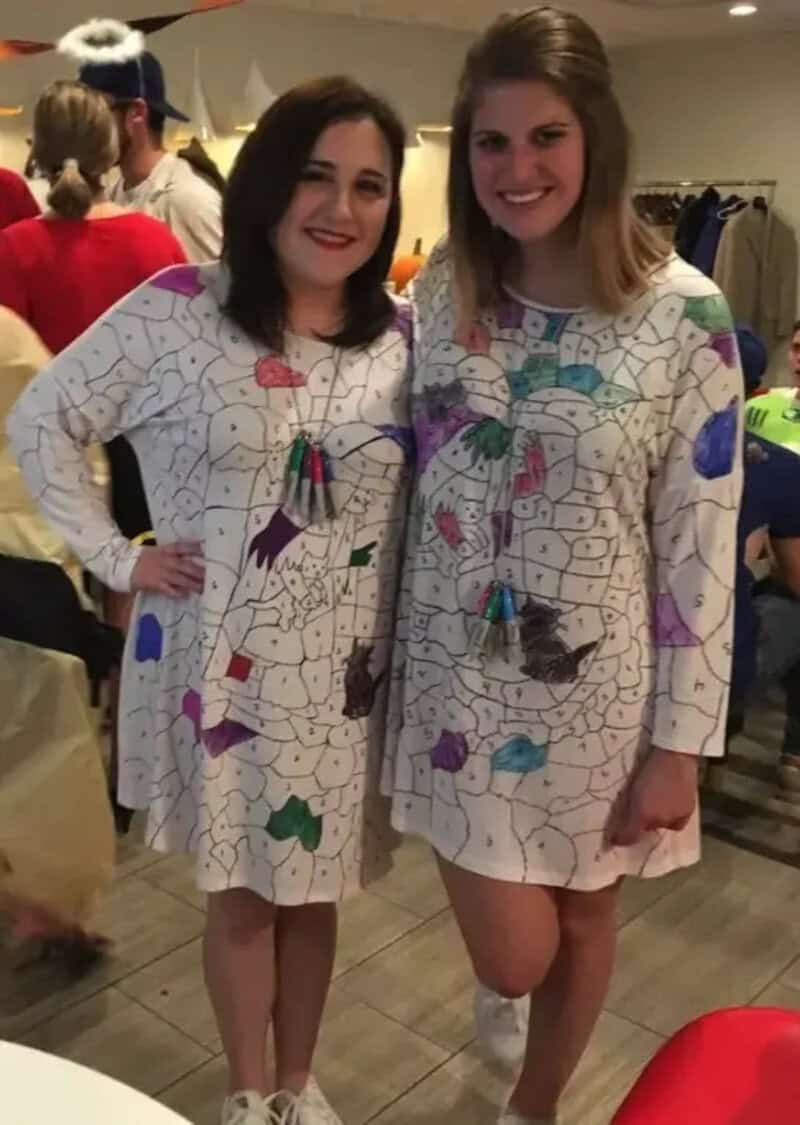 Two women in white long-sleeve dresses with colorful mosaic patterns pose together at a party.