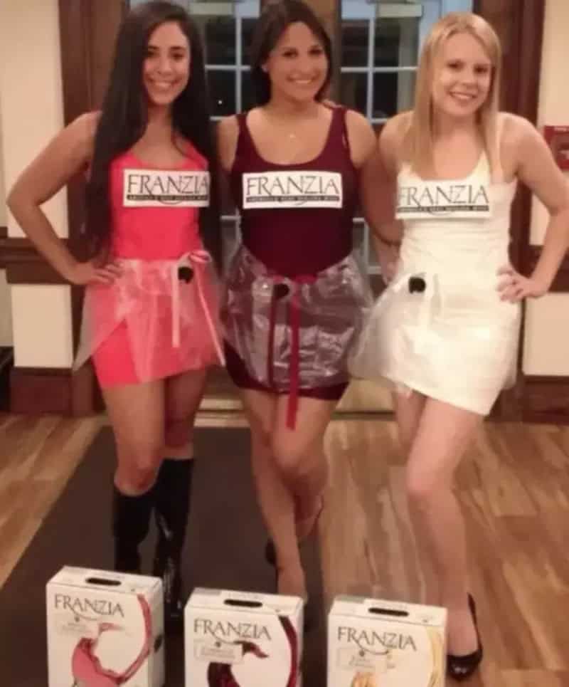 Three women in sexy outfits standing indoors with brand labels FRANZIA on their chests and wine gift.