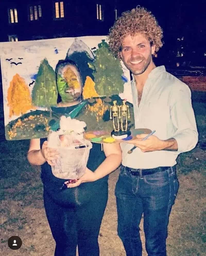Two people outdoors with Halloween makeup and costumes; one holds a painted board and a small bucket.