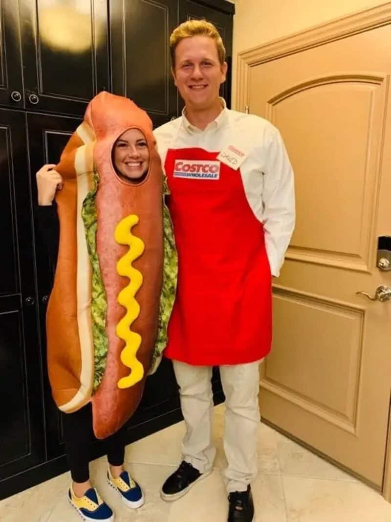 Two people posing in a kitchen; person on left wears a hot dog costume and blue sneakers, person on右.