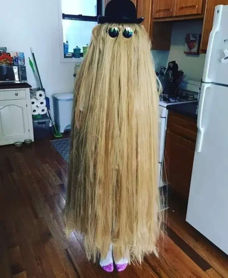 A person in a long blond wig with a black top hat and round sunglasses floats in a kitchen.