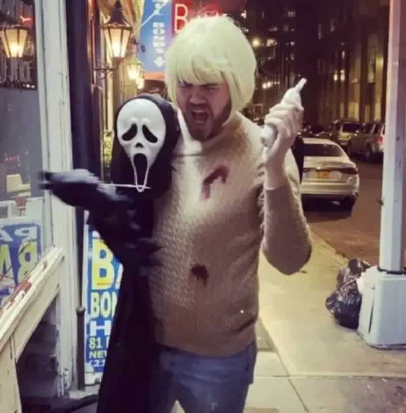 Man in beige sweater with blonde wig holding ghost-face puppet outside at night.