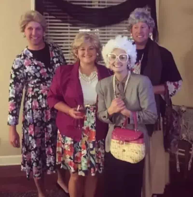 Group of four adults in retro outfits posing indoors with drinks; woman in red blazer, floral dress,.