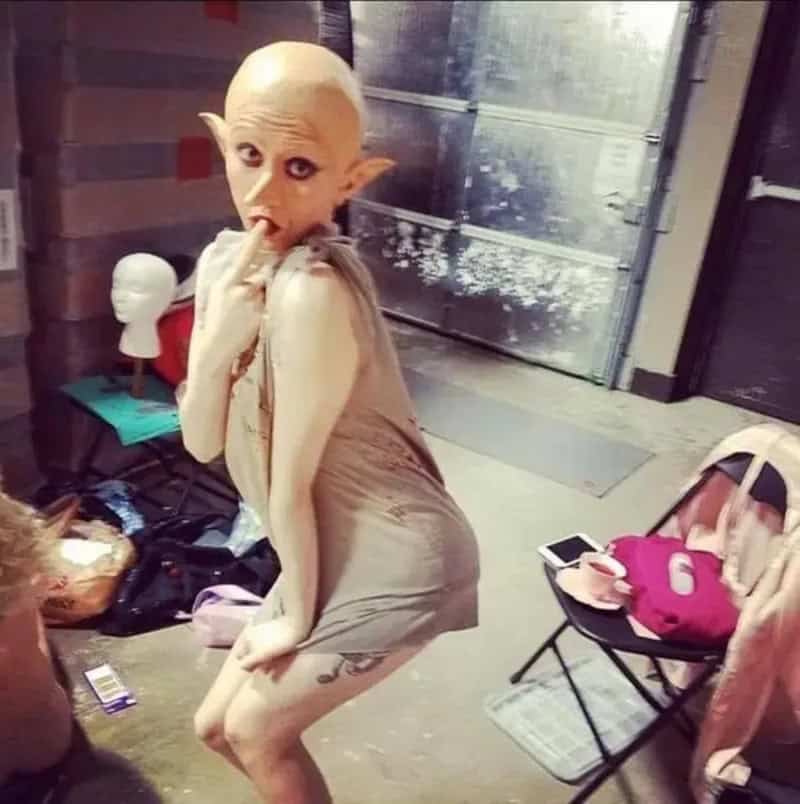 Front-facing alien-like figure with a bald head, wearing a tight beige outfit, standing in a clutter.