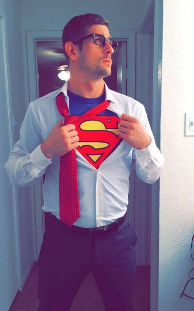 Man wearing white shirt and red tie pulling open shirt to reveal Superman logo on chest.