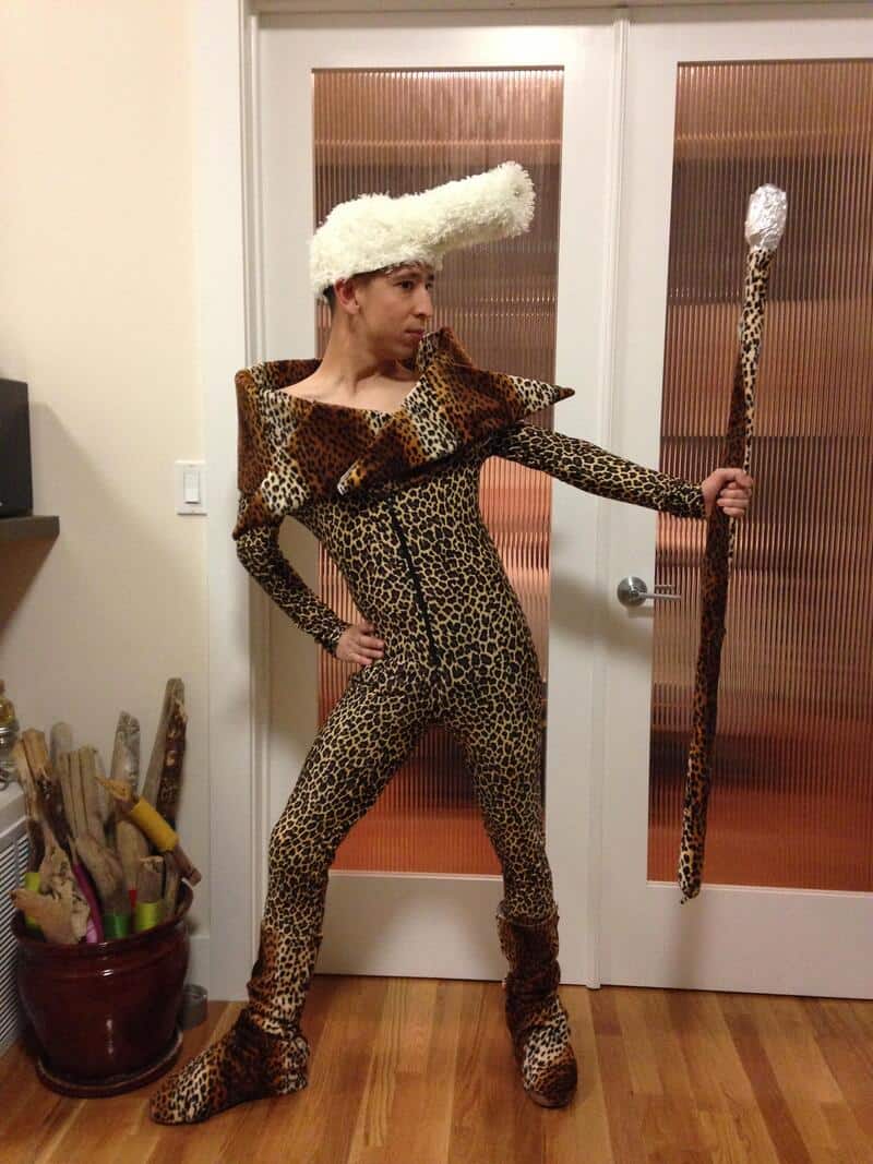 Person in leopard print outfit holding a staff with a pom-pom, standing in a doorway.