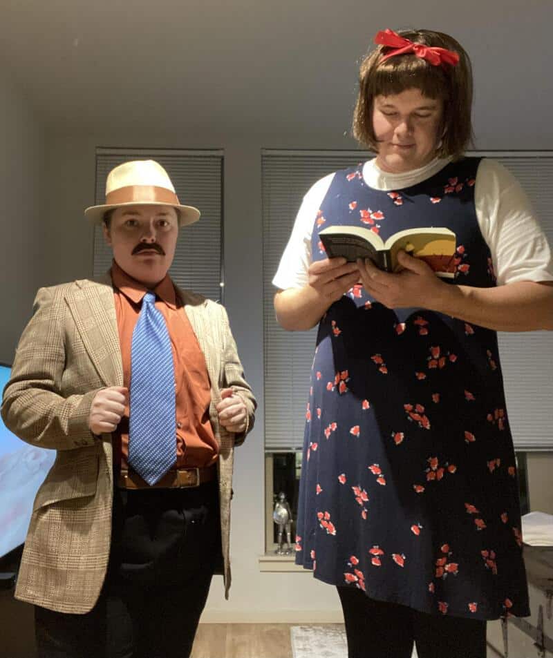 Two people in costumes: a man with a mustache wearing a tan fedora, brown blazer, orange shirt, and蓝.