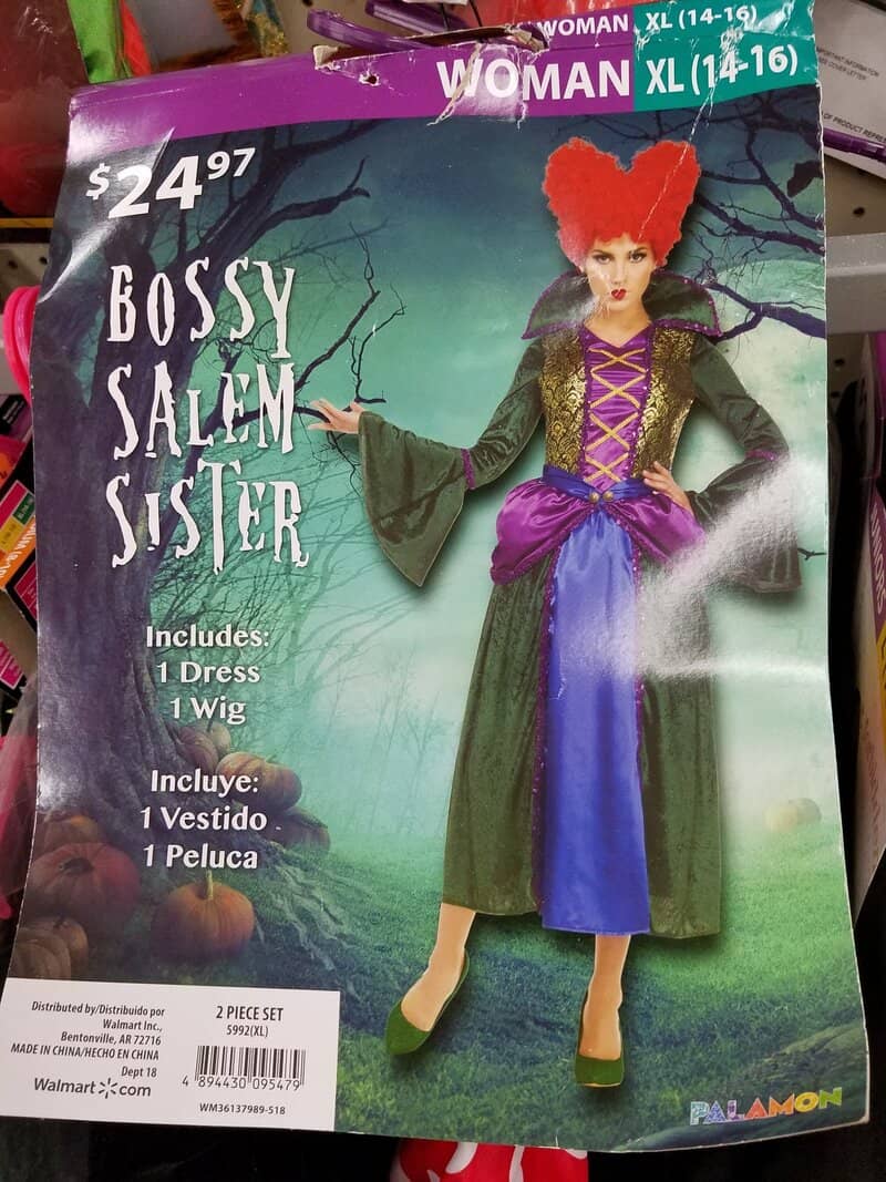 Costume of a witch with red hair, green dress, purple apron, and black cape, on Halloween-themed box.