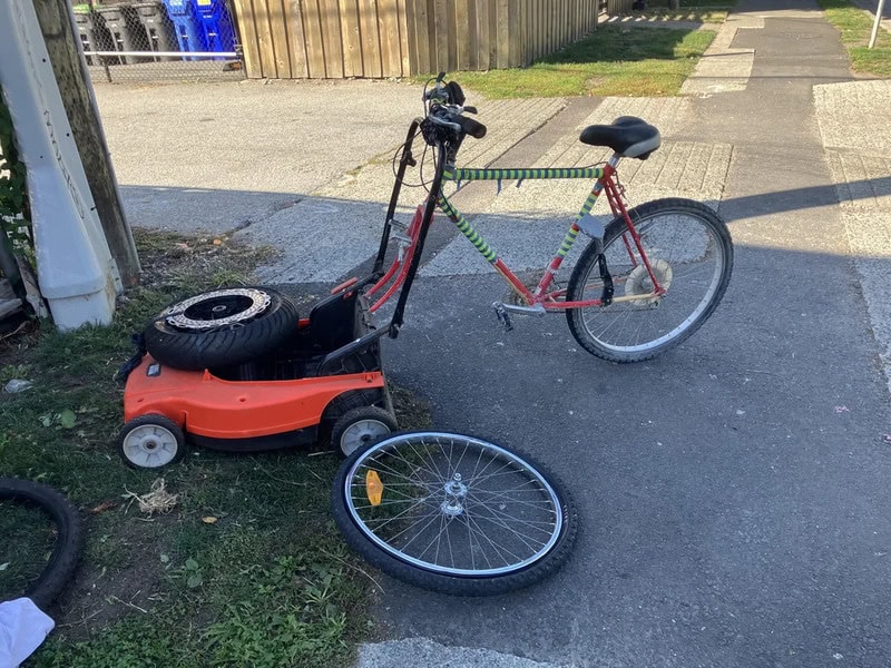 A red bicycle attached to a lawnmower with a detached wheel on the ground.