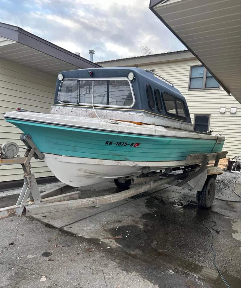 Small boat on trailer parked between two houses, under cloudy sky.