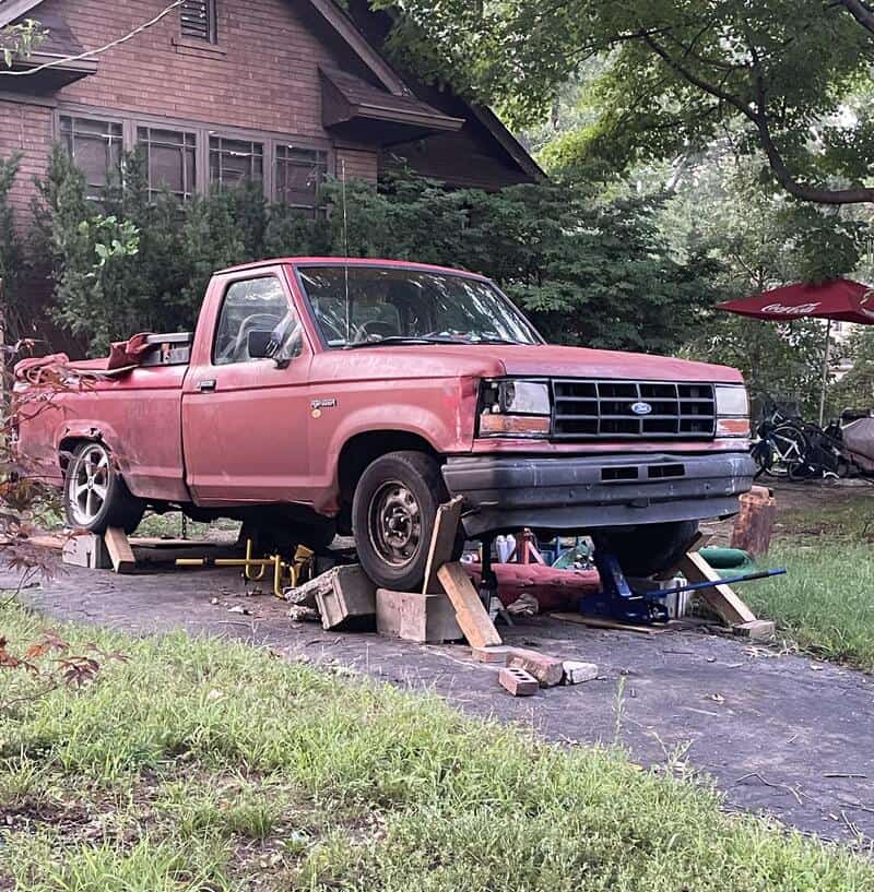 Pink Ford pickup truck on blocks with front wheels removed, supported by concrete blocks and wooden,.