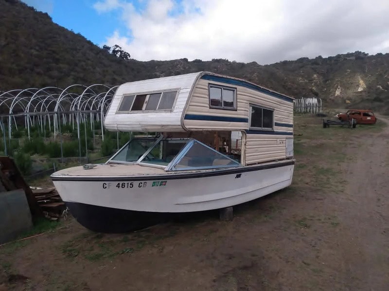 A boat with a camper on top, parked on dirt with mountains and a cloudy sky in the background.