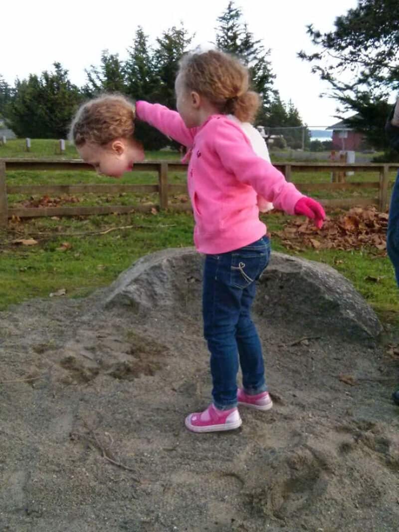 Girl in pink jacket balancing on a mound with a head and arms of another girl in the background.